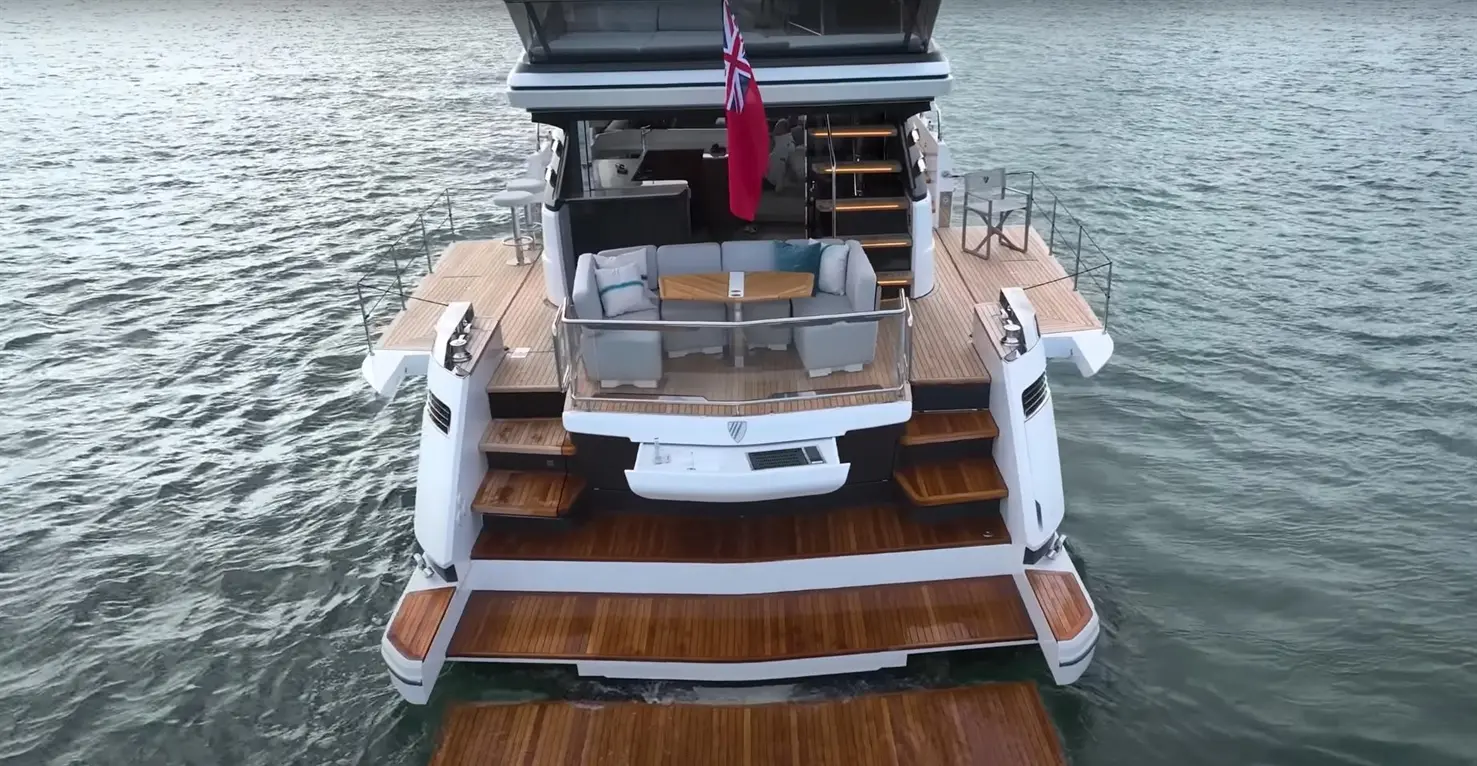 Luxury yacht with a wooden deck, a seating area on the stern, and a British flag flying above the cabin against water backdrop.